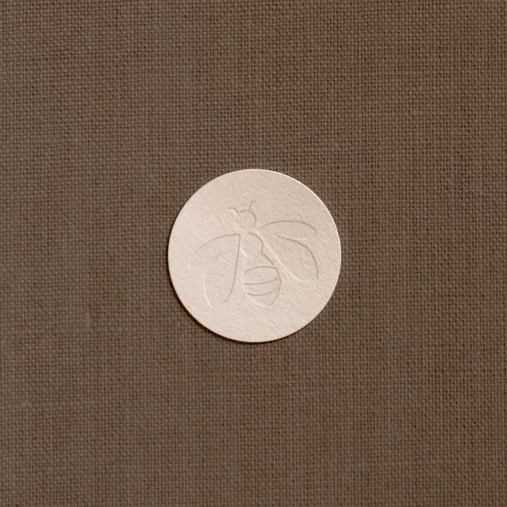 A letter-pressed mockup of a bee icon on soft pink paper, over a brown linen background.